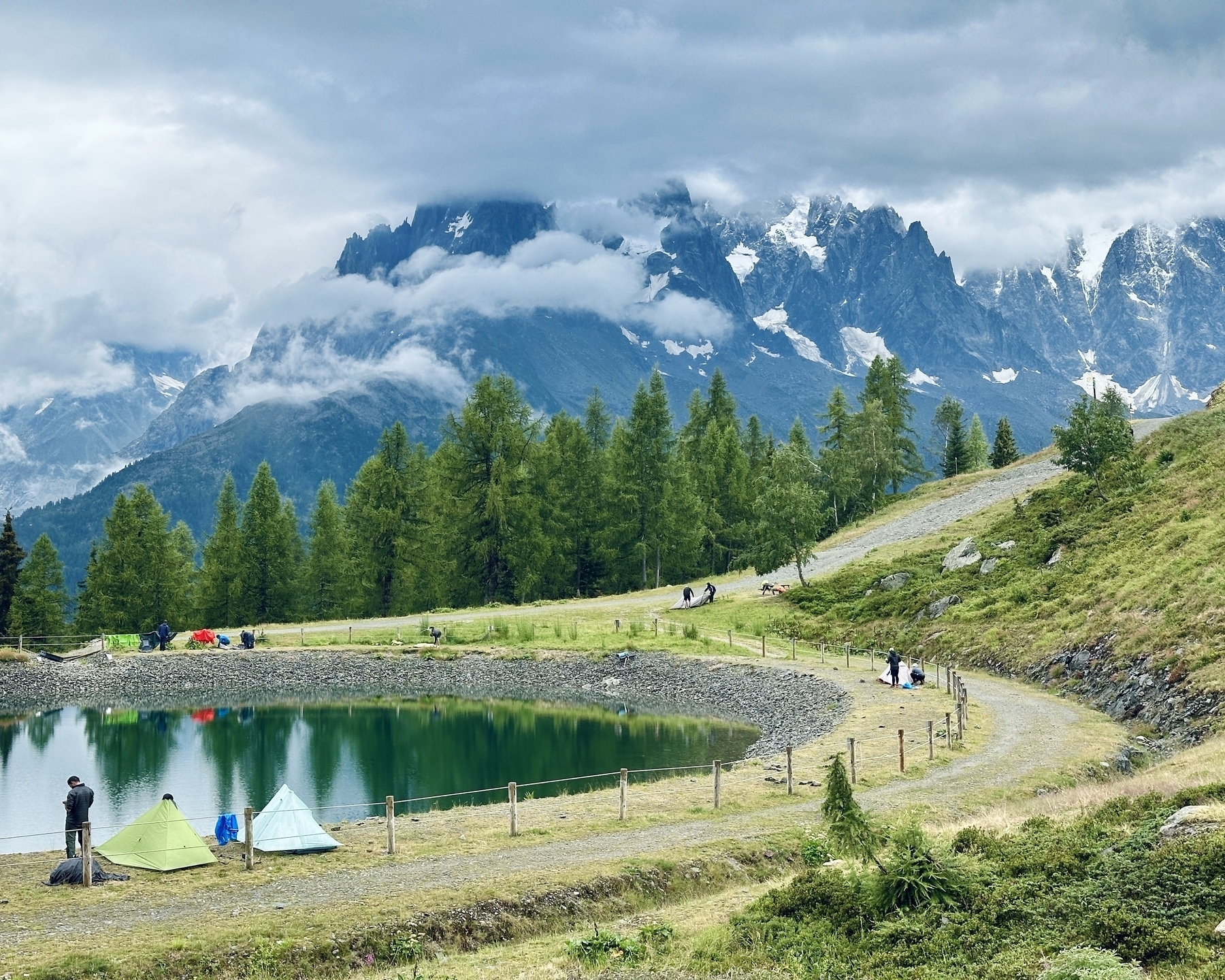 A tranquil landscape featuring tents set up by a small pond, surrounded by lush greenery and towering mountains under a cloudy sky.