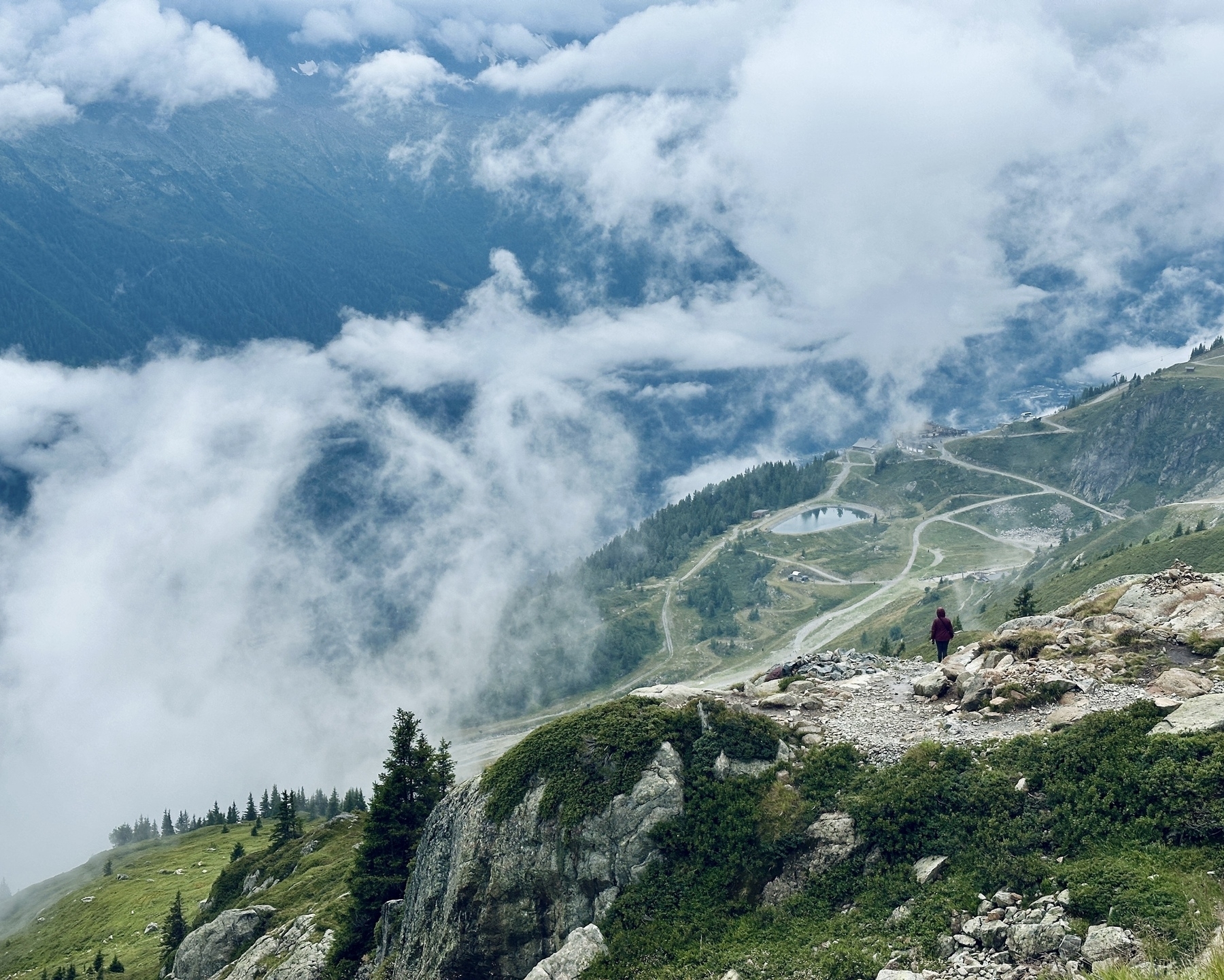 A lone hiker stands on a rocky mountain ridge overlooking a valley with winding paths and misty clouds.